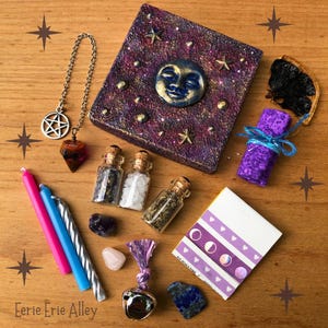 Könnte beinhalten: Eine Sammlung mystischer Gegenstände, darunter eine verzierte Schachtel mit einem Mond, Sternen und kleinen Glasflaschen mit Korken. Enthält auch Kerzen, ein Pendel, Kristalle und eine Glocke. Der Text "Eerie Erie Alley" ist sichtbar.
