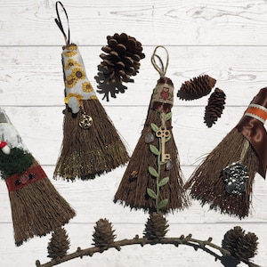 Pode incluir: Quatro vassouras em miniatura decoradas com vários enfeites, incluindo uma estampa de girassol, uma chave, uma pequena coruja e uma pedra preciosa vermelha. As vassouras são exibidas em um fundo de madeira branca com pinhas.