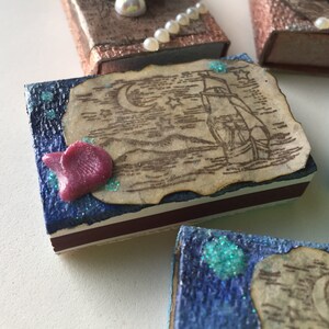 May include: A blue matchbox with a vintage nautical scene on the top. The scene features a ship sailing on the ocean under a crescent moon and stars. The matchbox has a pink resin embellishment and a white and brown border.
