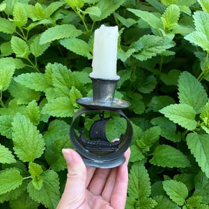 Pode incluir: Um castiçal metálico com base circular e um design central recortado de um navio viking com vela. O suporte sustenta uma vela cilíndrica branca e espessa que se encaixa dentro de um pequeno copo no topo da estrutura metálica.