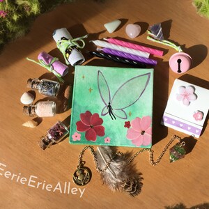 Pode incluir: Uma pintura em tela verde que retrata uma borboleta com flores rosa e vermelhas. A pintura é cercada por vários objetos, incluindo pequenas garrafas, velas, um sino, um cristal e um bloco de notas com um desenho de flor rosa. O texto "EerieErieAlley" é visível no canto inferior esquerdo da imagem.