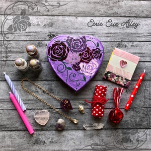 May include: A purple heart-shaped box with a floral design, three small glass bottles, a pink and white striped notebook, a pink heart-shaped crystal, a gold chain with a purple pendant, a white crystal, a red bell, a red and white polka dot wrapped roll, two pink candles, and a white candle with blue stripes. The text "Eerie Erie Alley" is written in black at the top of the image.