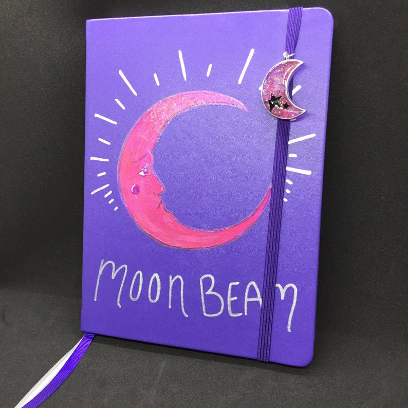 May include: Purple notebook with a pink crescent moon design and the words "Moon Beam" written in white. A silver charm with a crescent moon design is attached to the notebook.