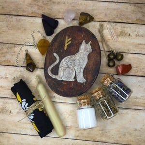 May include: An assortment of spiritual items, including a wooden cat carving, crystals, small glass bottles with corks, a candle, and a black cloth with a floral design. The items are arranged on a wooden surface.