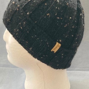 K&ouml;nnte beinhalten: Schwarze Strickm&uuml;tze mit einem melierten Muster und einem braunen Lederanh&auml;nger mit der Aufschrift "Tuscany".