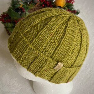 K&ouml;nnte beinhalten: Eine gr&uuml;ne Strickm&uuml;tze mit einem Zopfmuster und einem kleinen braunen Lederanh&auml;nger mit der Aufschrift "Weather".