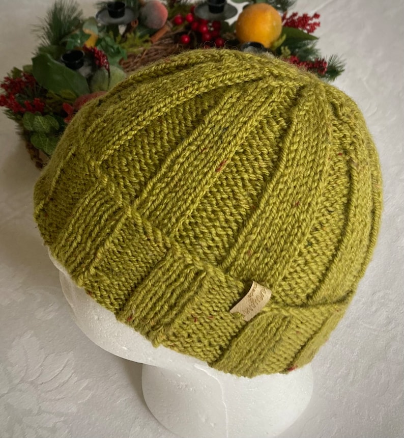 K&ouml;nnte beinhalten: Eine gr&uuml;ne Strickm&uuml;tze mit einem Zopfmuster und einem kleinen braunen Lederanh&auml;nger mit der Aufschrift "Weather".