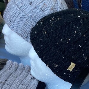 K&ouml;nnte beinhalten: Zwei Strickm&uuml;tzen auf Styropor-Mannequins. Die M&uuml;tze links ist hellgrau mit einer gesprenkelten Textur. Die M&uuml;tze rechts ist schwarz mit einer gesprenkelten Textur und einem braunen Etikett mit der Aufschrift "Tassle".