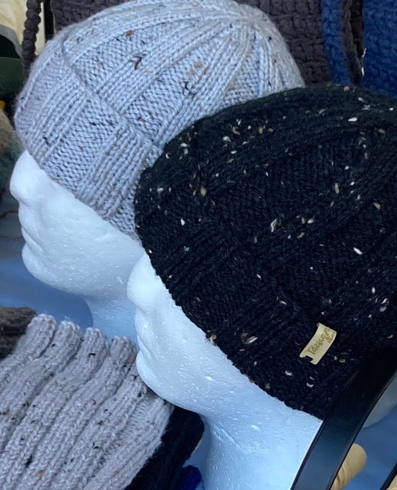 K&ouml;nnte beinhalten: Zwei Strickm&uuml;tzen auf Styropor-Mannequins. Die M&uuml;tze links ist hellgrau mit einer gesprenkelten Textur. Die M&uuml;tze rechts ist schwarz mit einer gesprenkelten Textur und einem braunen Etikett mit der Aufschrift "Tassle".