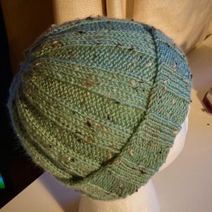 K&ouml;nnte beinhalten: Eine gr&uuml;ne und blaue Strickm&uuml;tze mit einem strukturierten Muster. Die M&uuml;tze sitzt auf einem wei&szlig;en Mannequin-Kopf.