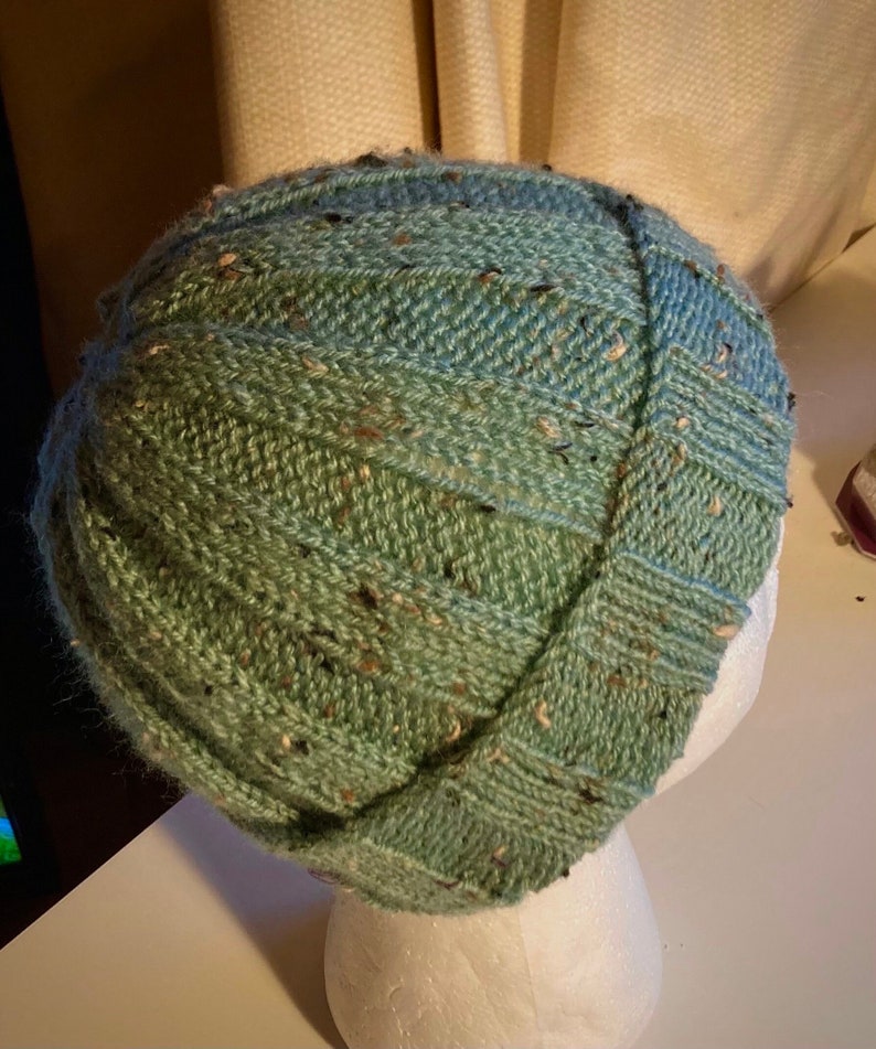 K&ouml;nnte beinhalten: Eine gr&uuml;ne und blaue Strickm&uuml;tze mit einem strukturierten Muster. Die M&uuml;tze sitzt auf einem wei&szlig;en Mannequin-Kopf.