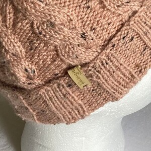 K&ouml;nnte beinhalten: Eine rosa Strickm&uuml;tze mit Zopfmuster und einem kleinen braunen Lederanh&auml;nger mit der Aufschrift "Handmade".