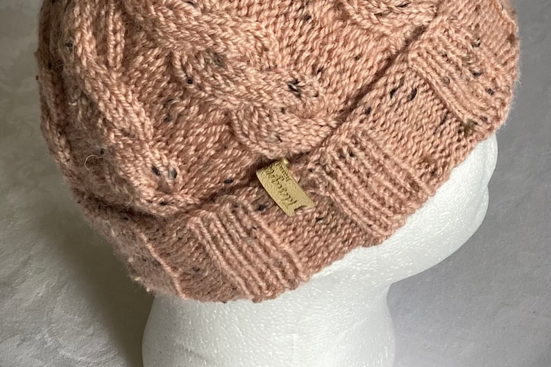 K&ouml;nnte beinhalten: Eine rosa Strickm&uuml;tze mit Zopfmuster und einem kleinen braunen Lederanh&auml;nger mit der Aufschrift "Handmade".
