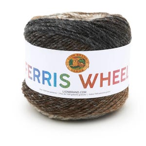 May include: A round skein of Lion Brand Ferris Wheel yarn featuring a variegated color palette of dark charcoal, brown, and cream tones. A white paper label wraps around the center, displaying the brand logo and the text FERRIS WHEEL.