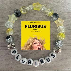 May include: A beaded bracelet with clear, yellow, and black beads, and star-shaped accents. The word "PLURIBUS" is spelled out in white letter beads. A yellow square with the text "PLURIBUS Volume 1" and a woman's face is in the center.