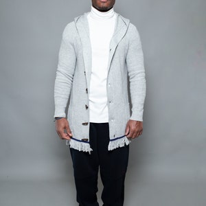 May include: A man wearing a gray hooded cardigan with a white turtleneck shirt and black pants. He is wearing gray and white sneakers.