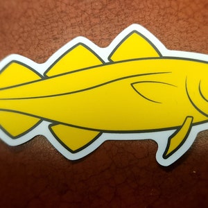 May include: A yellow fish sticker with a white border. The fish has a streamlined body with fins and a tail. The sticker is on a brown surface.