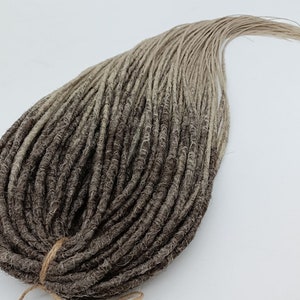 Könnte beinhalten: Ein Bündel synthetischer Dreadlocks in Braun- und Blondtönen. Die Dreadlocks sind eng gewellt und haben ein natürliches, strukturiertes Aussehen. Sie sind mit einer braunen Schnur zusammengebunden.