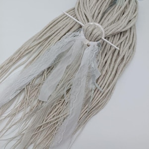 May include: Light gray dreadlocks with a white hair accessory. The hair accessory includes a white circular piece, a white stick, and white lace ribbons. A light blue ribbon is tied at the top of the dreadlocks.