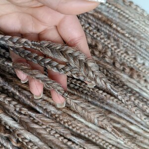 May include: A close-up of a bundle of brown and blonde synthetic dreadlocks. The dreadlocks are braided and have a natural, textured look.