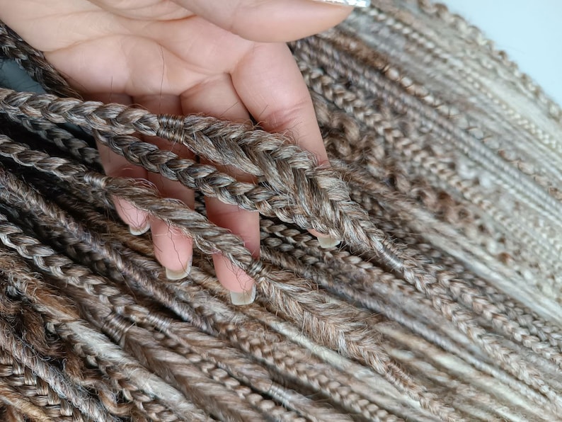May include: A close-up of a bundle of brown and blonde synthetic dreadlocks. The dreadlocks are braided and have a natural, textured look.