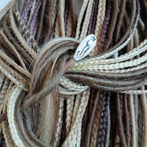 May include: A close-up of a bundle of synthetic dreadlocks in various shades of brown, blonde, and white. The dreadlocks are tied together with a wooden hair stick with a ceramic cat design.