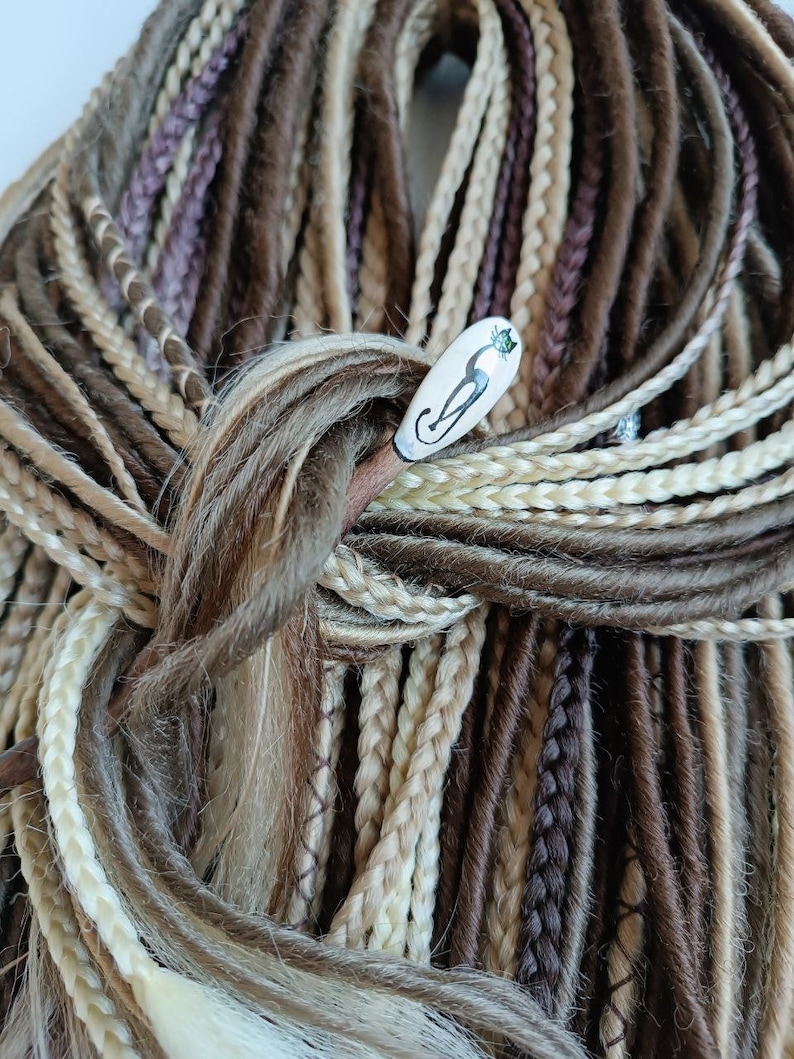 May include: A close-up of a bundle of synthetic dreadlocks in various shades of brown, blonde, and white. The dreadlocks are tied together with a wooden hair stick with a ceramic cat design.