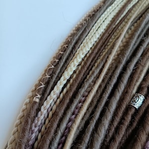 May include: A close-up of a bundle of synthetic dreadlocks in shades of brown, beige, and blonde. The dreadlocks are arranged in a neat, organised pattern, with some strands braided.