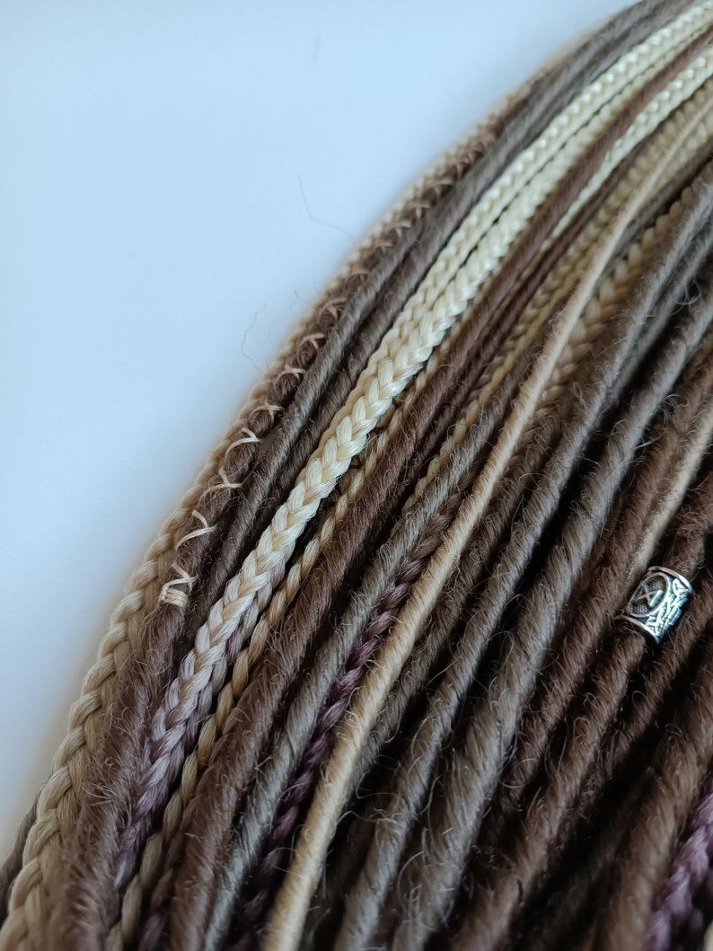 May include: A close-up of a bundle of synthetic dreadlocks in shades of brown, beige, and blonde. The dreadlocks are arranged in a neat, organised pattern, with some strands braided.