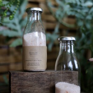 May include: Two glass bottles with silver lids, one labeled "Dandelion Organics Grapefruit Bath Salts." The label lists Epsom salts, Dead Sea salts, and pink Himalayan salt. The bottles are filled with pinkish bath salts, and sit on a wooden block.