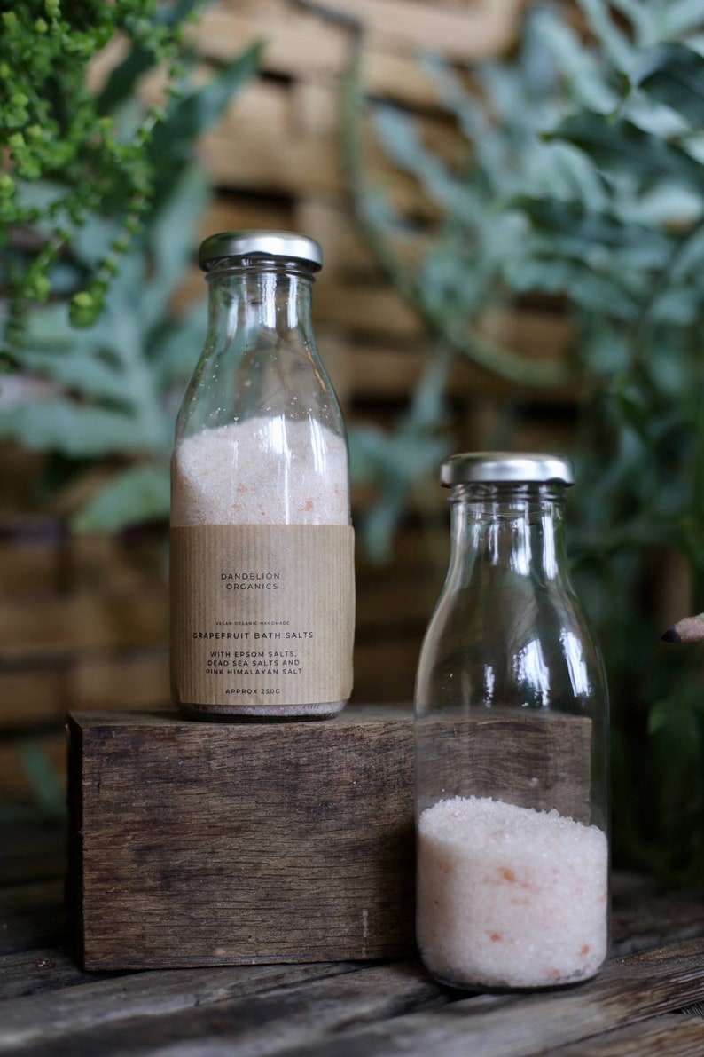 May include: Two glass bottles with silver lids, one labeled "Dandelion Organics Grapefruit Bath Salts." The label lists Epsom salts, Dead Sea salts, and pink Himalayan salt. The bottles are filled with pinkish bath salts, and sit on a wooden block.