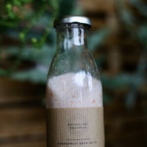 May include: A clear glass bottle with a silver cap, filled with pink bath salts. The label reads "Dandelion Organics" and "Grapefruit Bath Salts." The bottle contains approximately 250g of salts, including Epsom, Dead Sea, and Pink Himalayan salts.