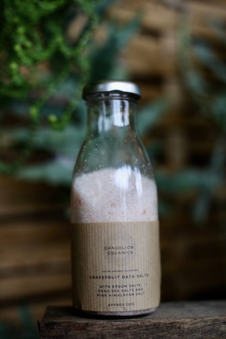 May include: A clear glass bottle with a silver cap, filled with pink bath salts. The label reads "Dandelion Organics" and "Grapefruit Bath Salts." The bottle contains approximately 250g of salts, including Epsom, Dead Sea, and Pink Himalayan salts.