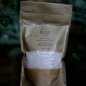 May include: A brown paper bag of orange bath salts. The bag has a clear window showing the pinkish salt crystals. Text on the bag reads "Dandelion Organics" and "Orange Bath Salts" with Epsom, Dead Sea, and Pink Himalayan salts. Approximately 250g.