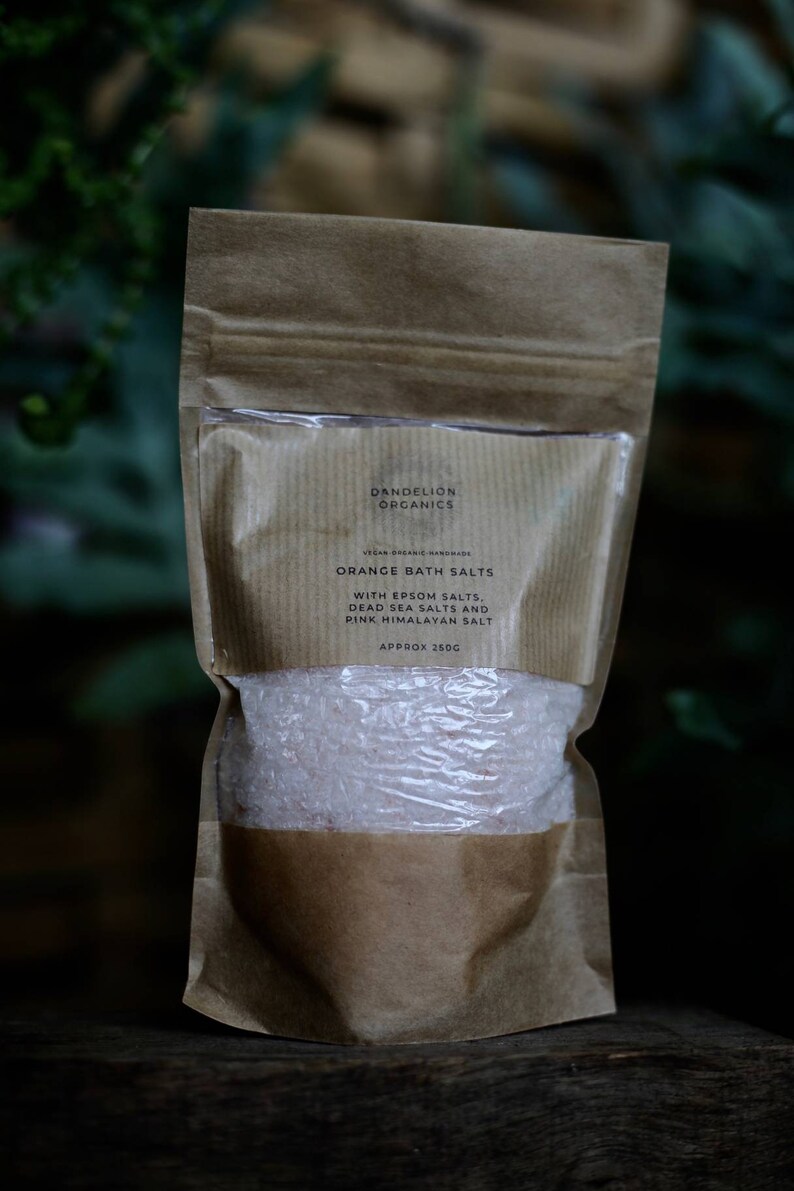 May include: A brown paper bag of orange bath salts. The bag has a clear window showing the pinkish salt crystals. Text on the bag reads "Dandelion Organics" and "Orange Bath Salts" with Epsom, Dead Sea, and Pink Himalayan salts. Approximately 250g.