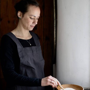 May include: A woman wearing a dark gray apron and black long-sleeved shirt, standing near a window. She is holding a wooden spoon over a wooden bowl filled with a white substance. The background includes a wooden door and a white wall.