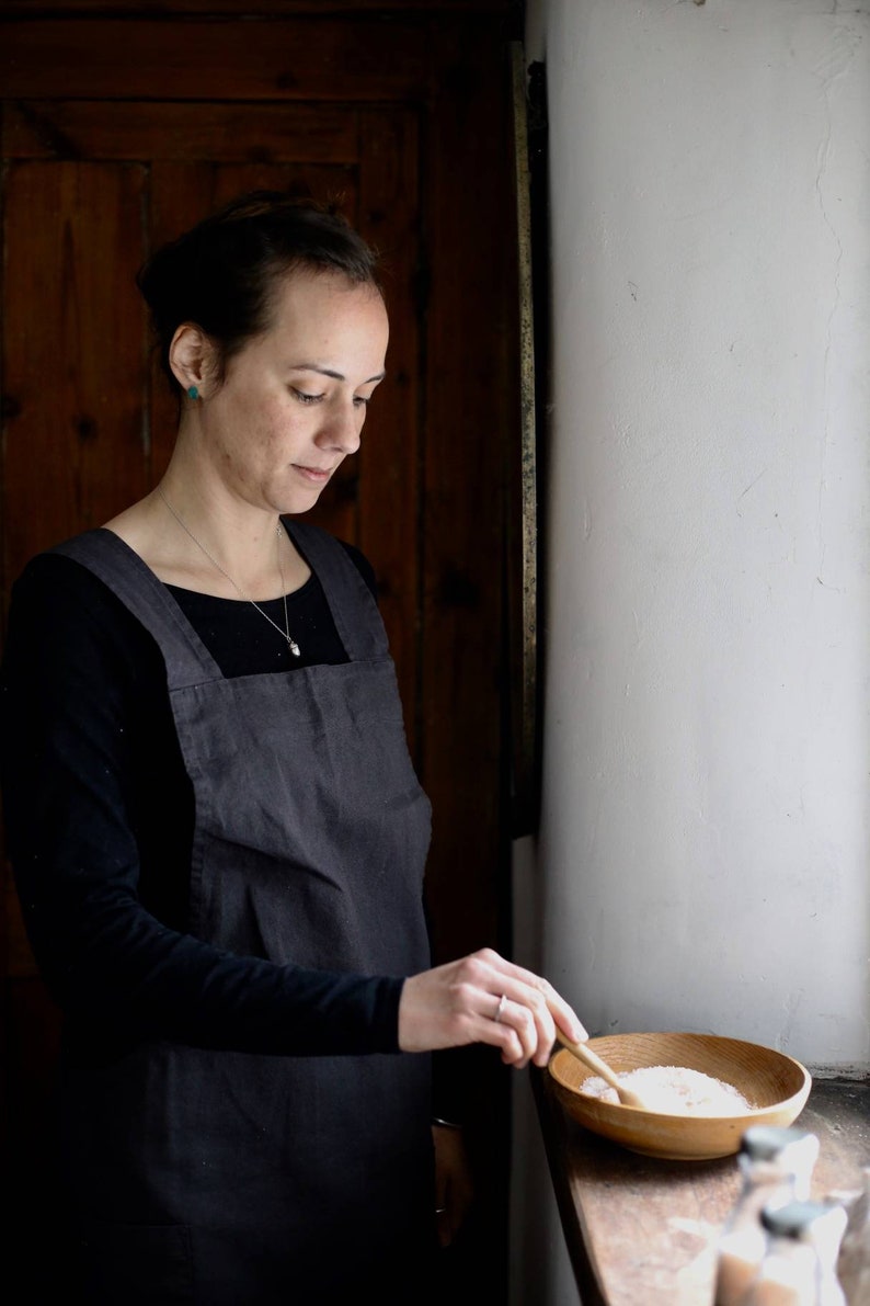 May include: A woman wearing a dark gray apron and black long-sleeved shirt, standing near a window. She is holding a wooden spoon over a wooden bowl filled with a white substance. The background includes a wooden door and a white wall.