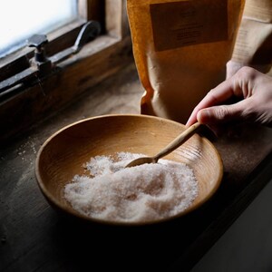 May include: A wooden bowl filled with coarse pink salt, with a wooden spoon resting in the salt. The bowl sits on a wooden surface near a window. A hand is holding the spoon. A brown paper bag is in the background.