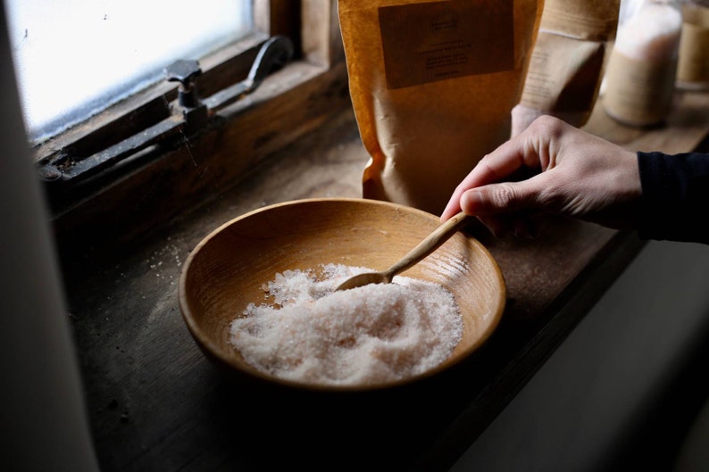 May include: A wooden bowl filled with coarse pink salt, with a wooden spoon resting in the salt. The bowl sits on a wooden surface near a window. A hand is holding the spoon. A brown paper bag is in the background.