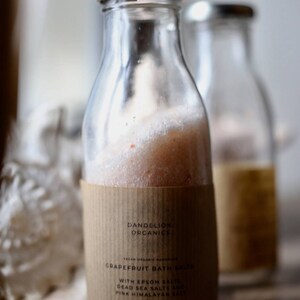 May include: Clear glass bottle filled with pink grapefruit bath salts, with a silver lid and a brown paper label. The label reads "Dandelion Organics" and "Grapefruit Bath Salts". The bottle contains approximately 250g of bath salts.