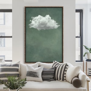 Puede incluir: Una impresión artística enmarcada que presenta una nube blanca y esponjosa sobre un fondo verde apagado. La obra de arte se exhibe sobre un sofá blanco con varias almohadas decorativas en tonos grises y blancos. La habitación tiene ventanas grandes y una estética minimalista.