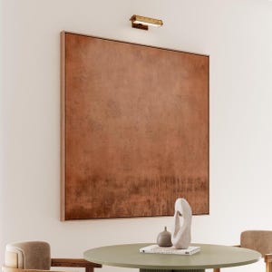 May include: A large, framed abstract painting with a textured, reddish-brown surface dominates the image. A small, gold-toned light fixture is mounted above the artwork. A round table with decorative objects sits below the painting.