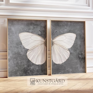 Peut inclure: Deux impressions d'art encadrées représentant des ailes de papillon blanches sur un fond gris texturé. L'œuvre est exposée dans une pièce avec des moulures blanches et un sol en bois. Le bas du cadre porte le texte "KUNSTGÅRD curated modern & vintage art prints."