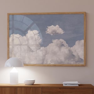 May include: A framed print depicting a tranquil sky with soft white clouds set against a pale blue background. The artwork is enclosed in a light brown wooden frame. A white lamp and bowl are on a wooden cabinet below.