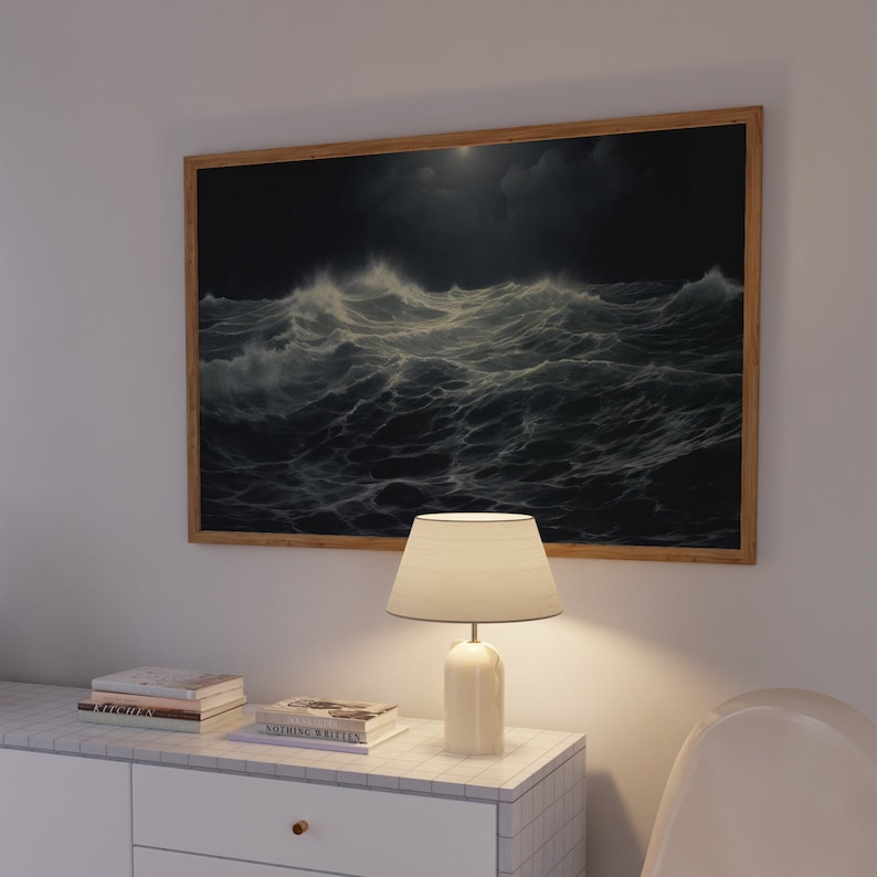 K&ouml;nnte beinhalten: Ein gerahmtes Gem&auml;lde eines st&uuml;rmischen Meeres bei Nacht mit einem Vollmond, der durch die Wolken scheint. Eine wei&szlig;e Tischlampe mit einem cremefarbenen Schirm steht auf einer wei&szlig;en Kommode mit einem Stapel B&uuml;cher.