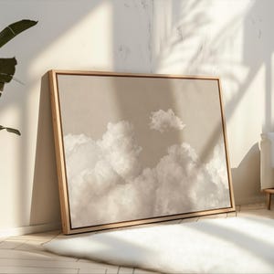 Könnte beinhalten: Ein gerahmter Leinwanddruck mit beigefarbenem Hintergrund und einem weißen Wolkenmuster. Der Druck ist in einem Holzrahmen und lehnt an einer weißen Wand.