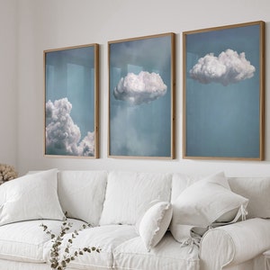 May include: Three framed prints of clouds against a blue sky. The prints are in wooden frames and are hung on a white wall above a white sofa.