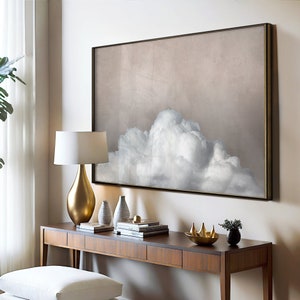 May include: A gold-framed canvas print of a large, white cloud against a light brown background. The print is hanging above a wooden console table with a gold lamp, decorative vases, and a gold metal sculpture.