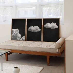 May include: Three black and white framed prints of clouds against a dark background. The prints are hanging on a wall above a light brown couch.