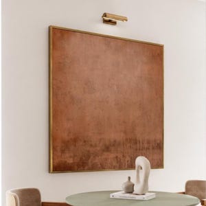 Könnte beinhalten: Ein großes quadratisches Wandbild mit einer texturierten, gesprenkelten Oberfläche in Kupfer- und Rosttönen, eingefasst in einen dünnen goldenen Rahmen. Über dem Kunstwerk ist eine kleine horizontale goldene Leuchte an der Wand montiert.